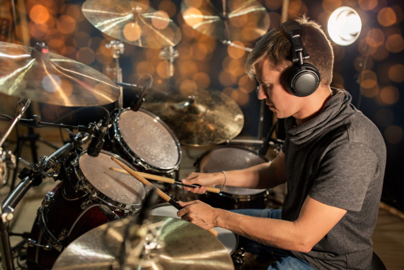 music, people, musical instruments and entertainment concept - male musician or drummer in headphones with drumsticks playing drums and cymbals at concert or studio over holidays lights background