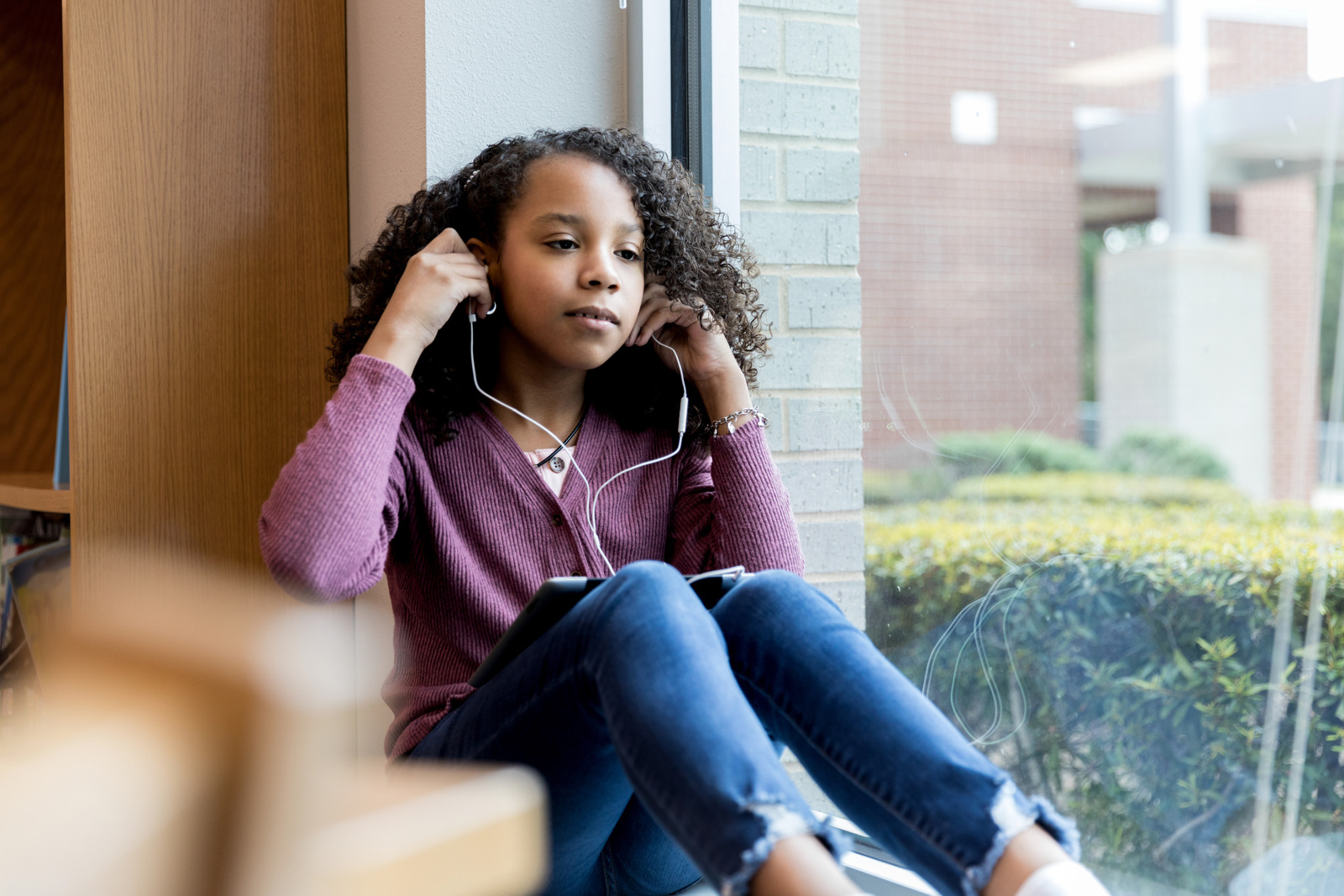 Taking a break from studying, the young sits on the library windowsill and adjusts her earbuds to listen to music on her digital tablet.