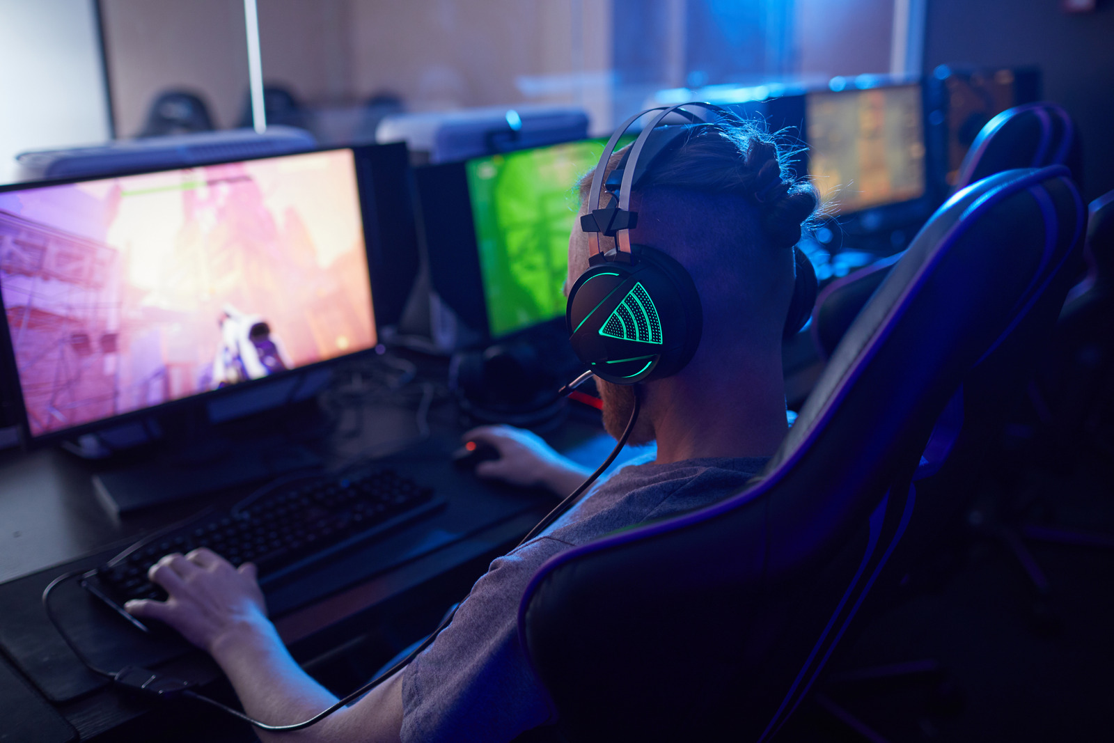 Rear view of young gamer wearing gaming headphones with backlight and playing in computer video game on computer in dark computer class