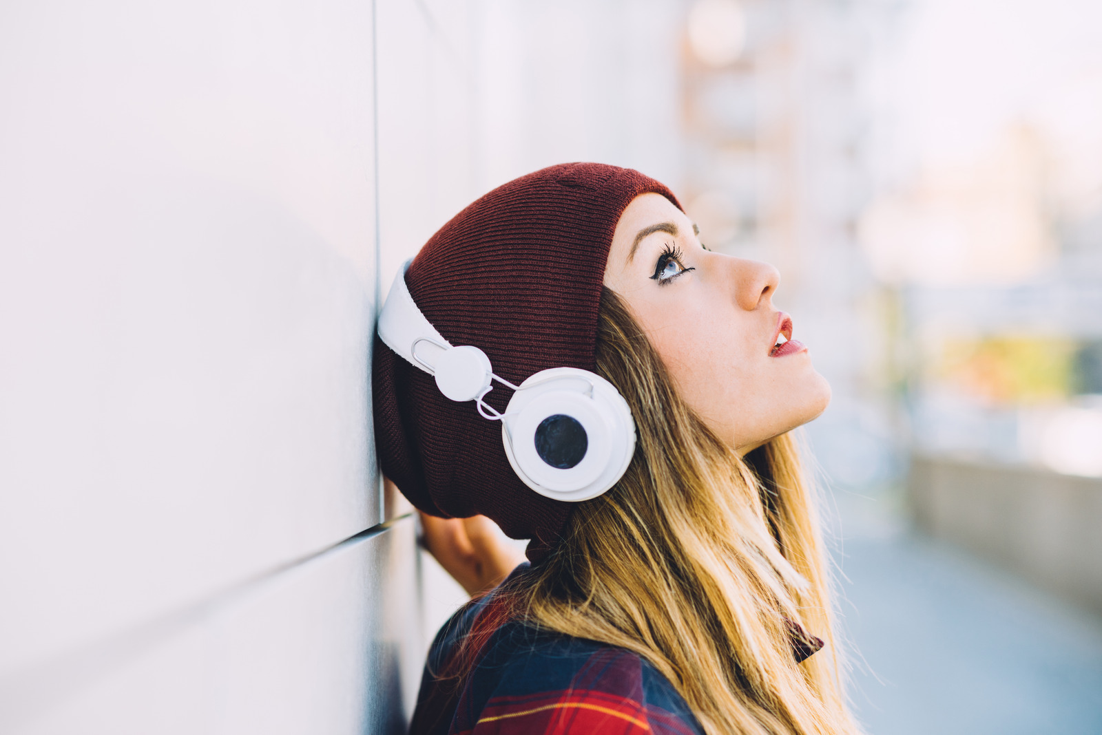 Half length profile portrait of young handsome caucasian blonde hair woman leaning against a wall, listening music with headphones, looking up - serene, enjoying, music concept