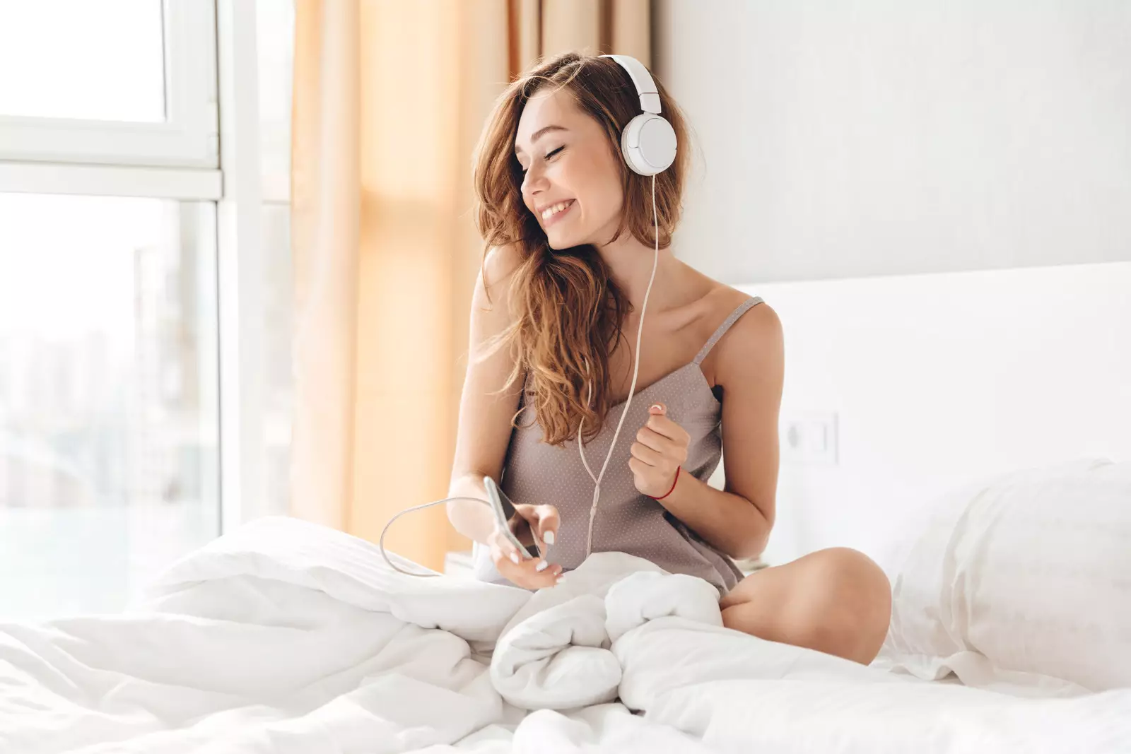 Carefree cheerful woman listening music on smartphone and wearing headphones in bed at home